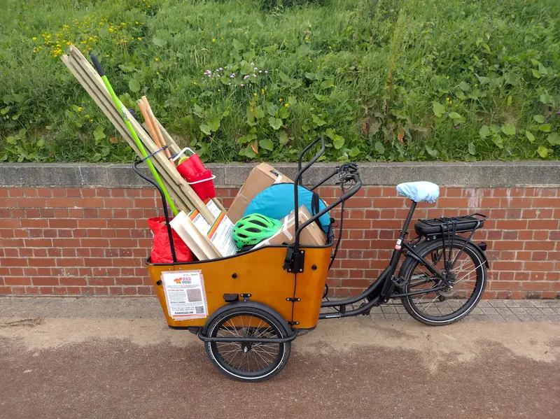A cargo tricycle packed with boxes, bags, a sandcastle bucket, brooms, rakes, and several long pieces of wood.