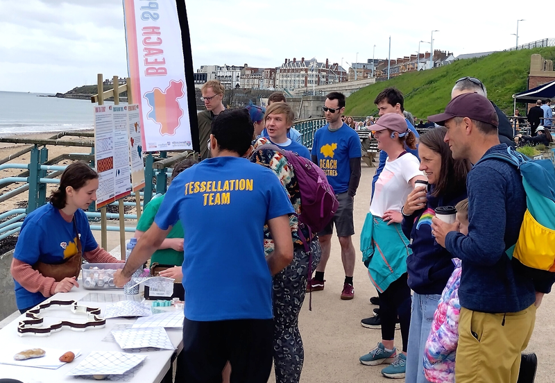 12 people stood in front of the information table on the prom. Volunteers are pointing at printed tessellations and 3D-printed shape cutters on the table.