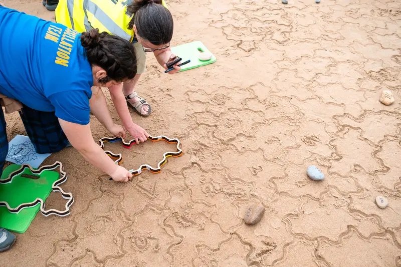 Two people fitting a Spectre stamper next to Spectre impressions in the sand.