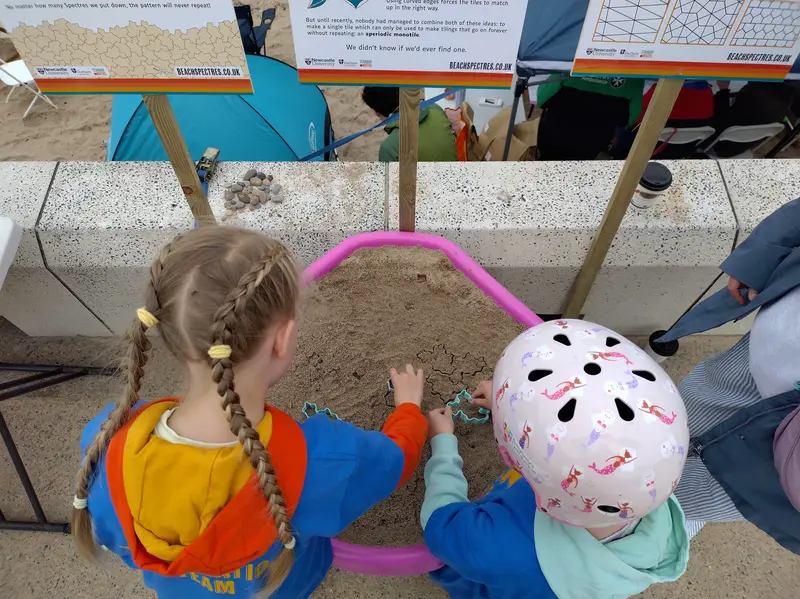 Looking down at the tuff tray. Two children are using the cutters to make impressions in the sand.