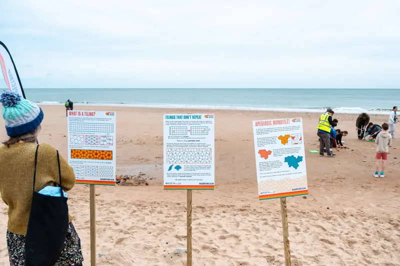 A person looking at the poster boards on the sand, with people working on the tiling in the background.