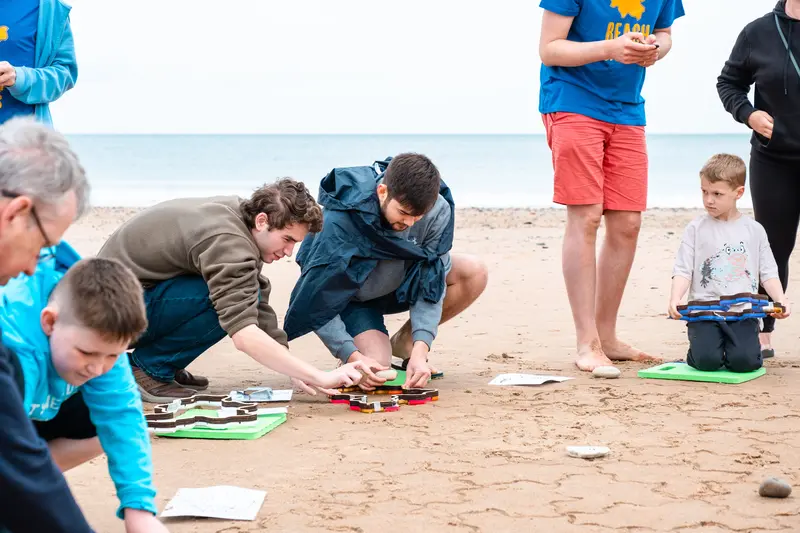 Two people working together to press a stamper into the sand, while a child holding a stamper looks on.