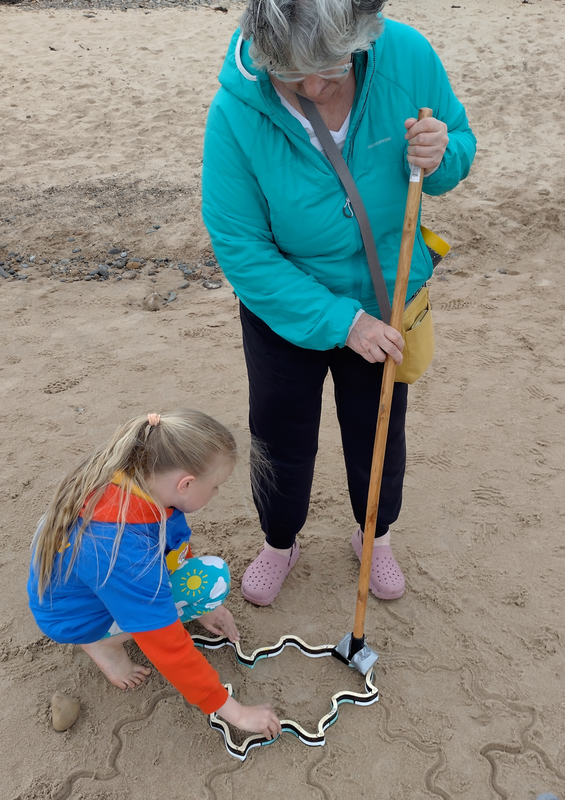 A grey-haired woman holding a broom handle and using it to tap a Spectre stamper, which is being held still by a blonde child.