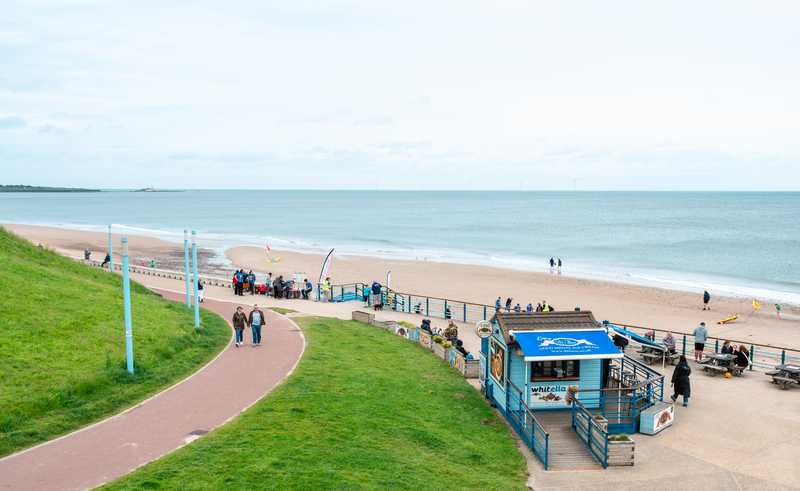 Looking down towards Whitley Bay prom and beach. An ice cream kiosk is at the end of the prom. A banner and information stand are at the top of the stairs leading down to the beach.