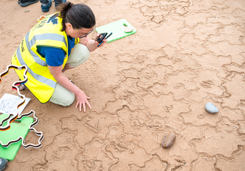 A person in a hi-viz kneeling and looing carefully at Spectre tiles which have had numbers drawn on the sand inside them.